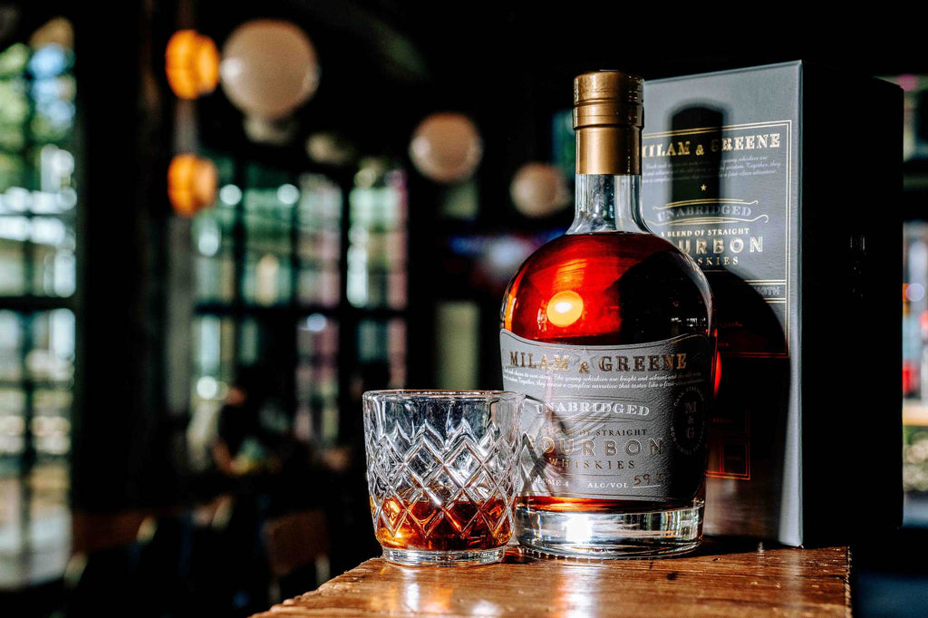 Bottle of Hill & Greene bourbon with a glass of whiskey on a wooden surface, blurred bar background