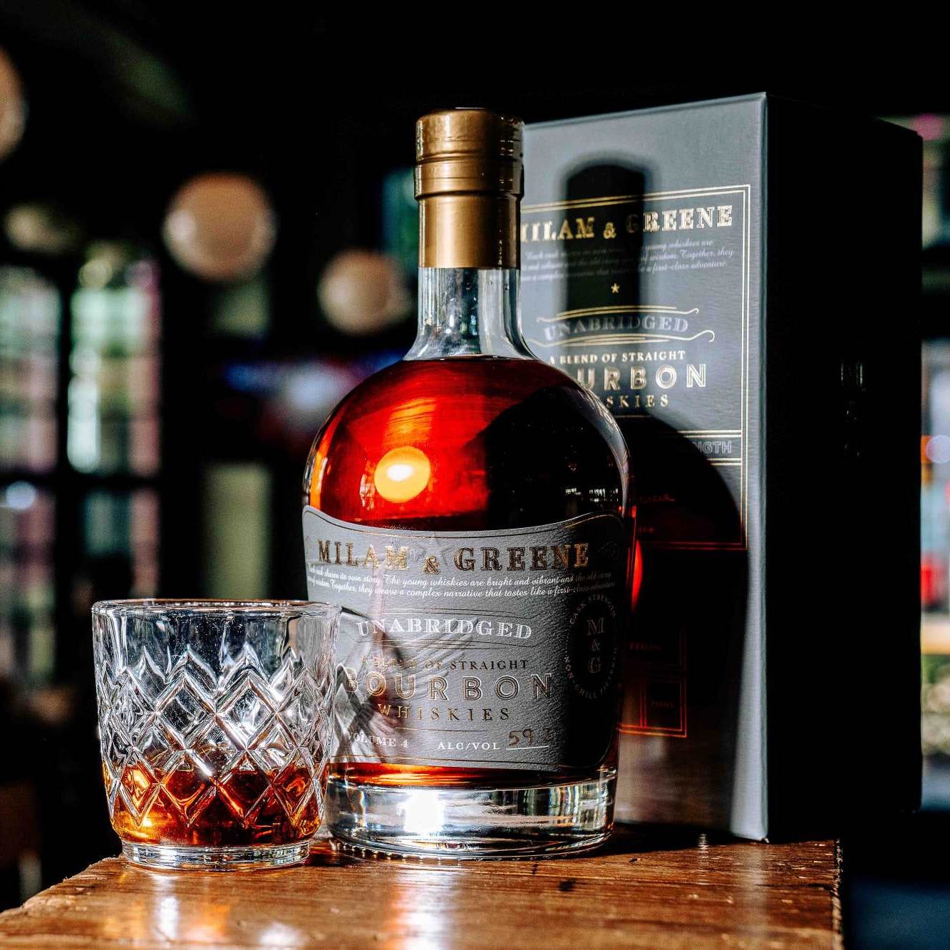 Bottle of Hill & Greene bourbon with a glass of whiskey on a wooden surface, blurred bar background