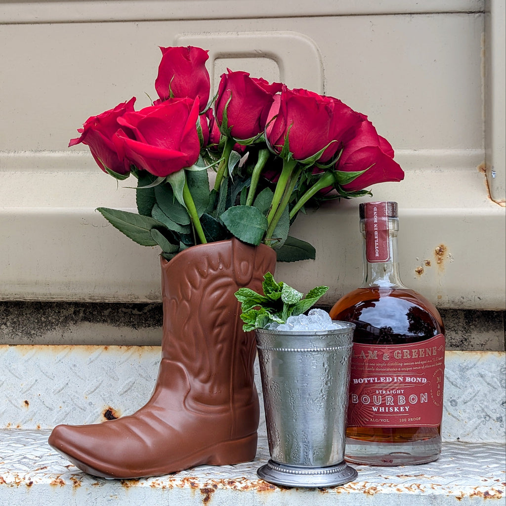 Red roses in a cowboy boot vase with a bottle of Jim Beam bourbon on a rustic surface.
