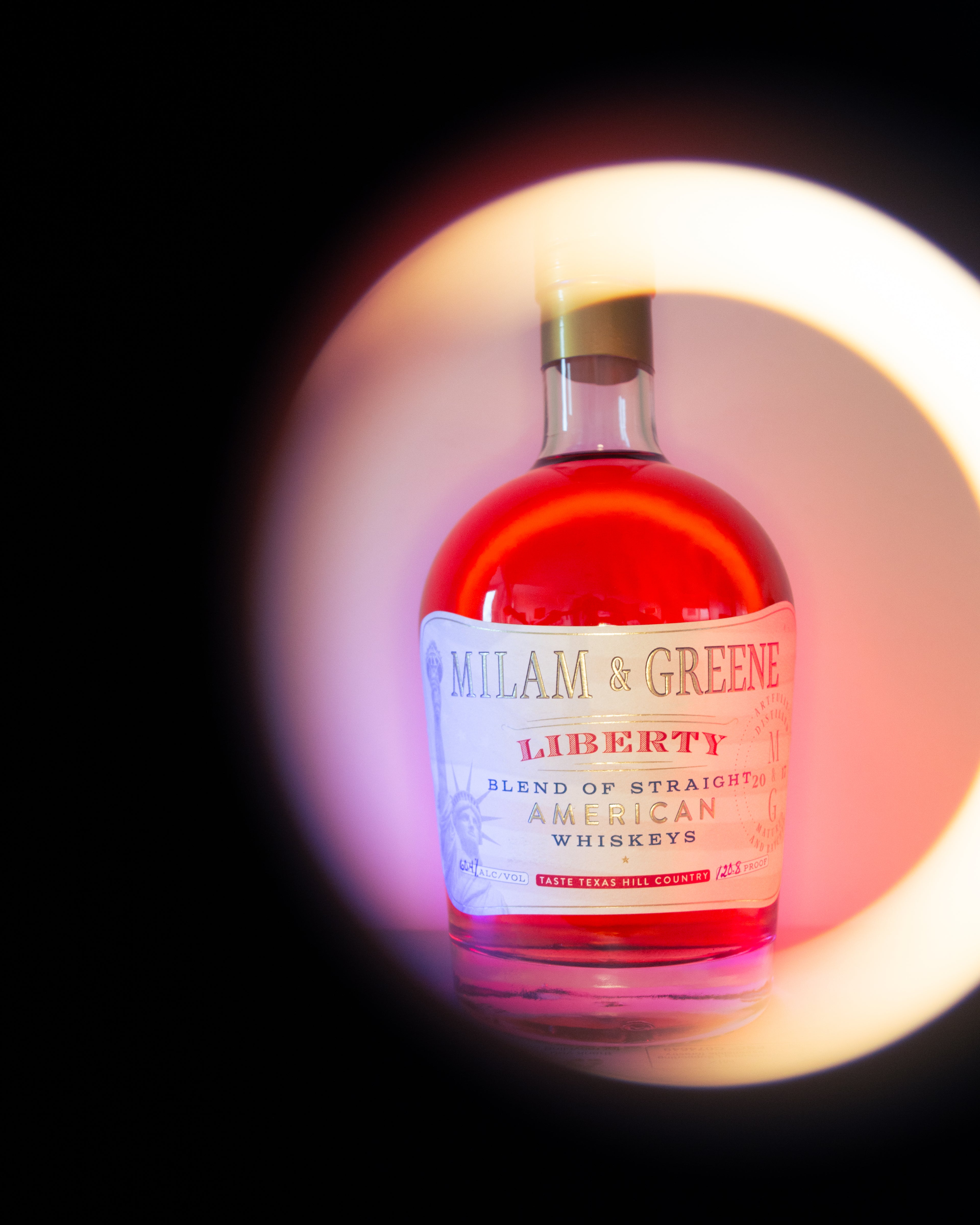 Bottle of Milam & Greene Liberty American Whiskey with a bright brown liquid inside, against a dark background.