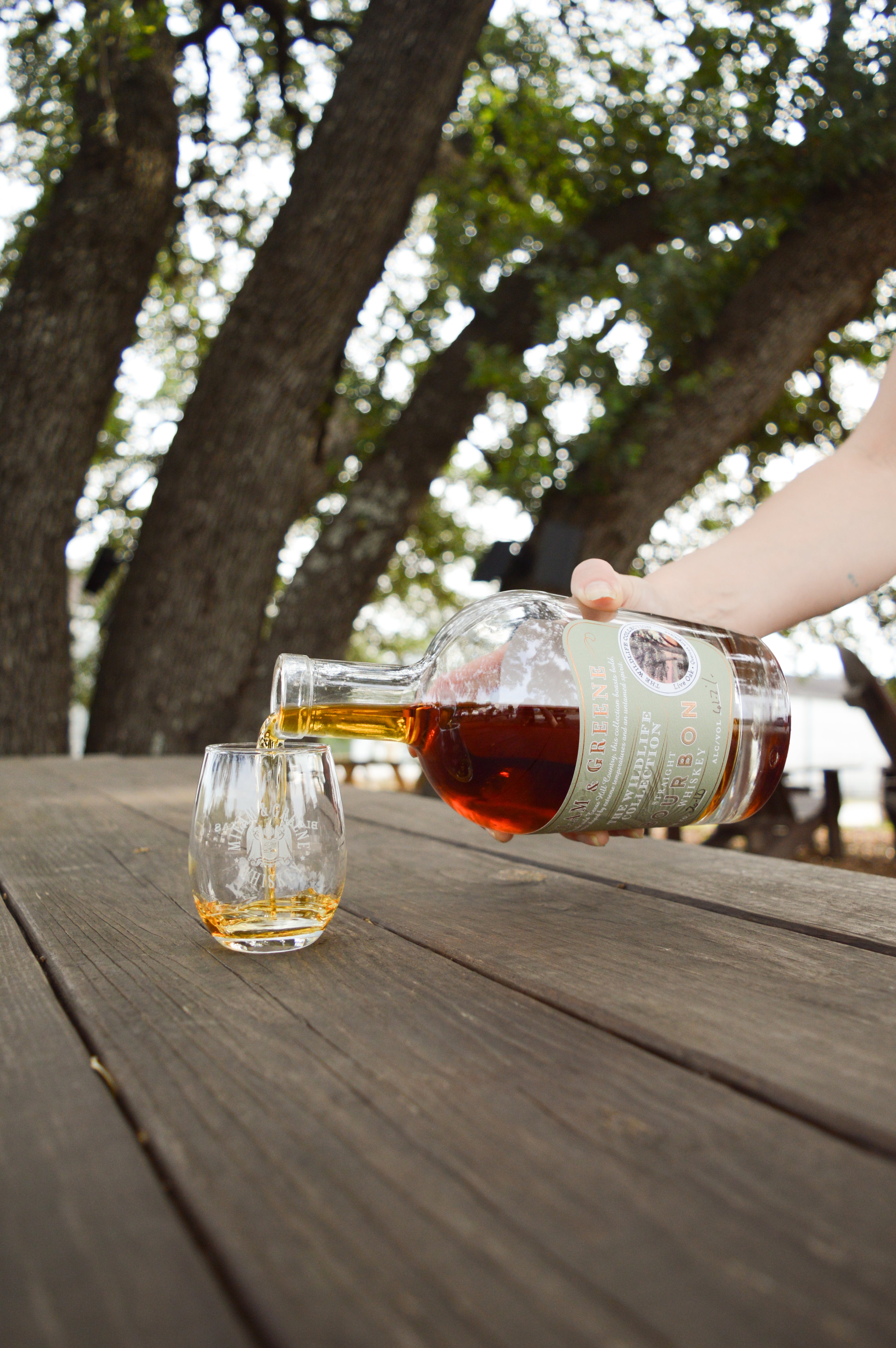 Milam and Greene Wildlife Collection Live Oak Straight Bourbon bottle of whiskey pouring into a glass on a picnic table under an oak tree