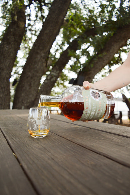 Milam and Greene Wildlife Collection Live Oak Straight Bourbon bottle of whiskey pouring into a glass on a picnic table under an oak tree