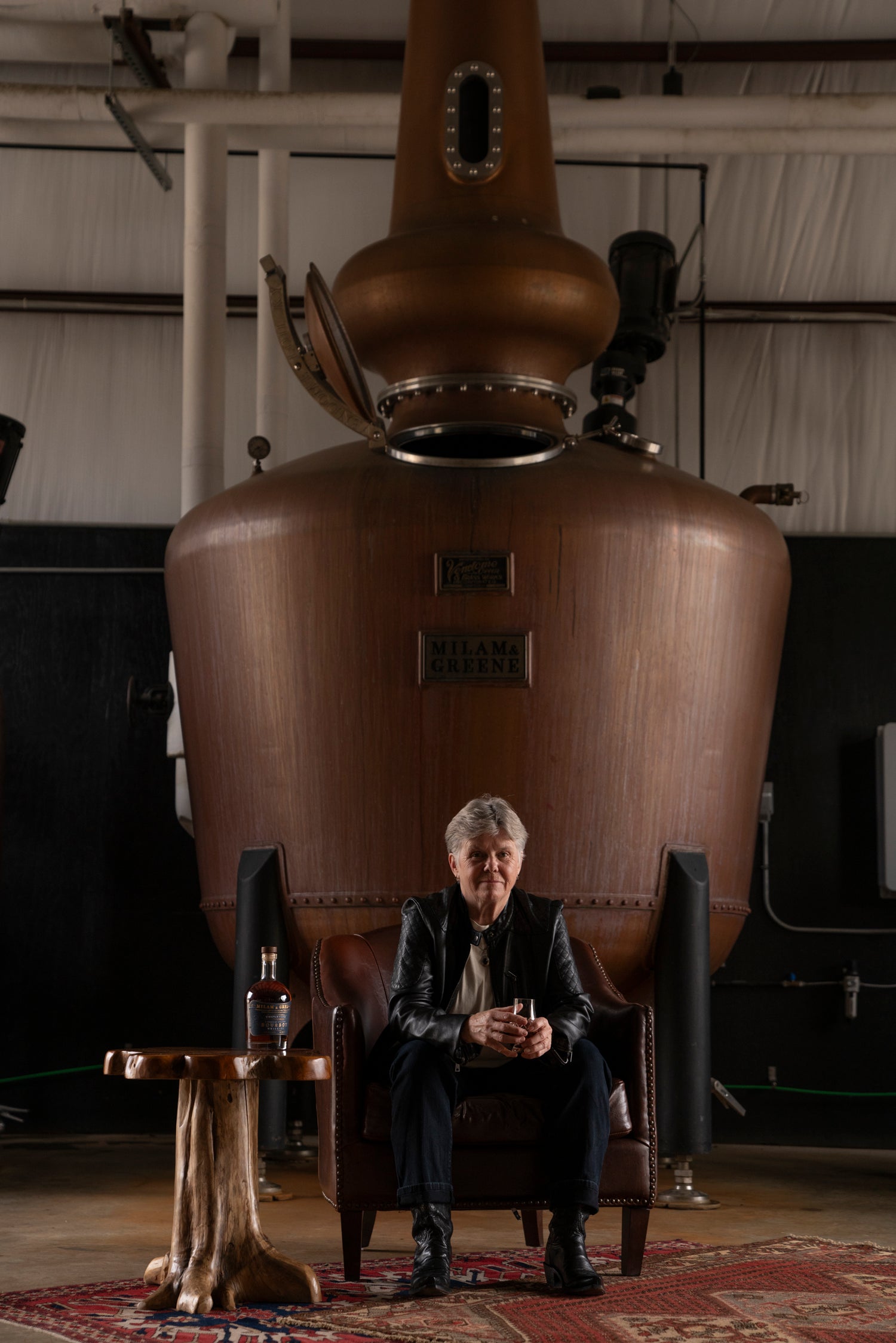 Marlene Holmes seated in front of the Milam & Greene 1000 gallon pot still with a bottle of Triple cask on a table next to her. 