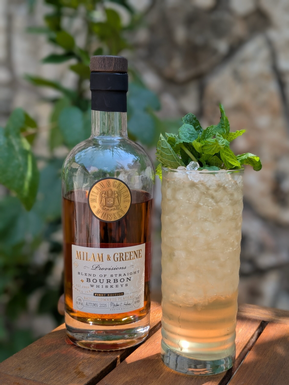 Bottle of Malt & Greene bourbon next to a cocktail with mint leaves on a wooden surface.
