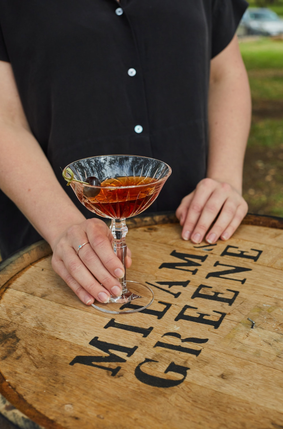 Person holding a cocktail glass on a wooden barrel with 'Milam & Greene' branding.