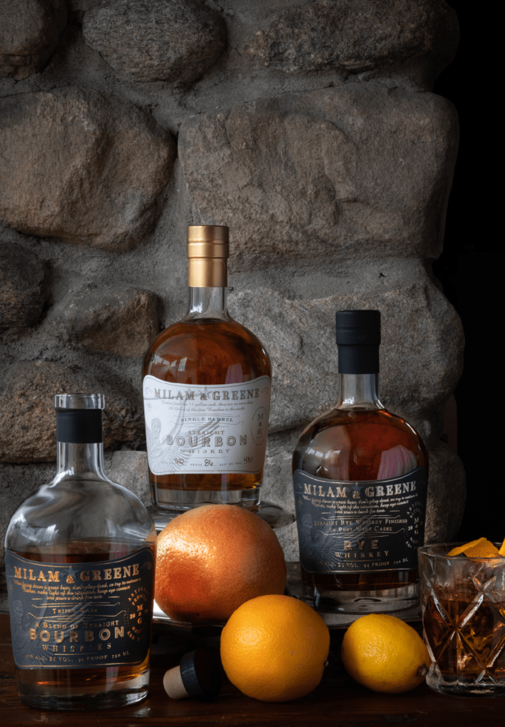 Three bottles of Milam & Greene bourbon with fruits and a glass on a stone surface.