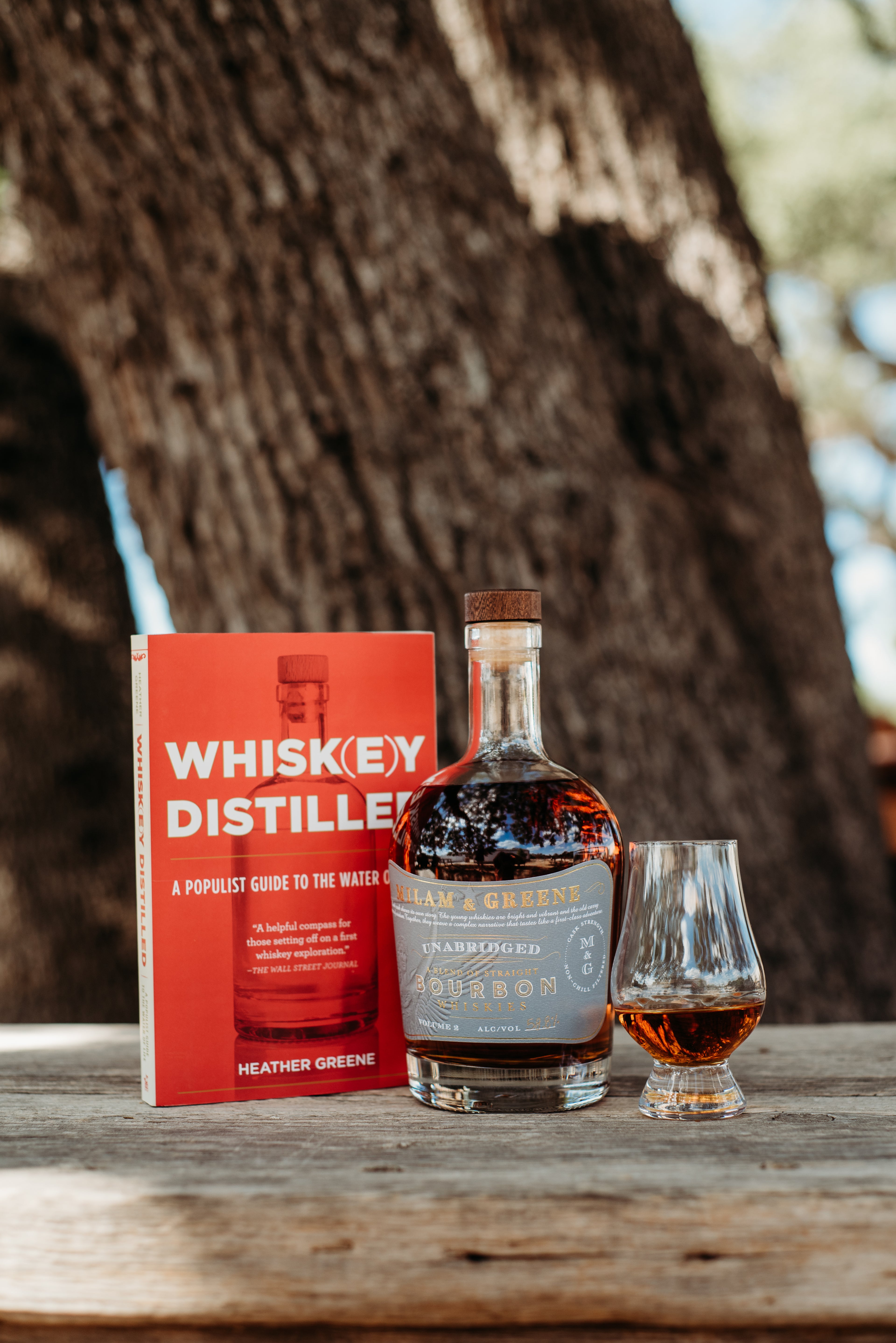 bottle of bourbon with a grey label in between a red book and a glass of neat whiskey in front of a tree