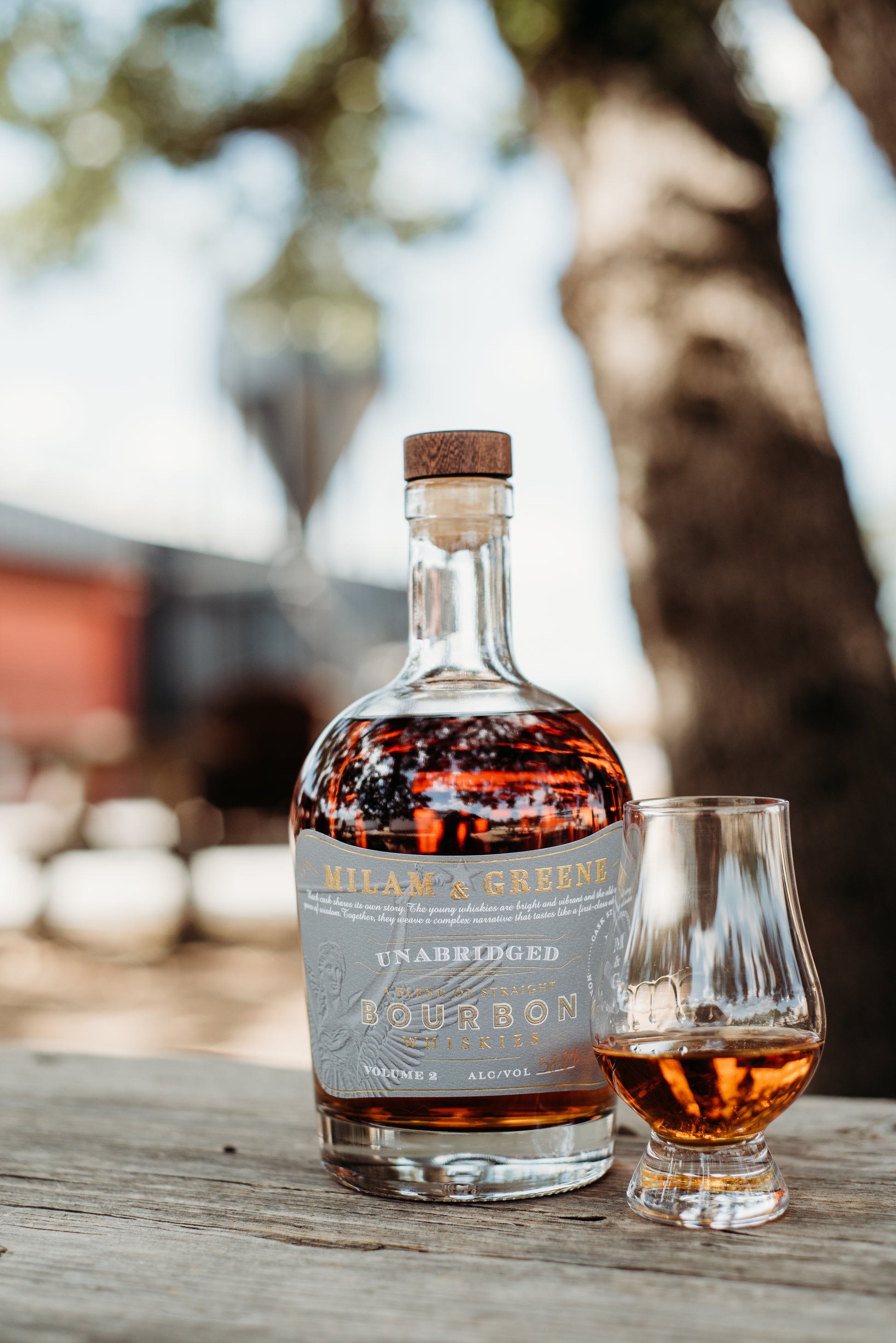 bourbon bottle with a grey label and a glass of poured whiskey in a glencairn in front of the milam and greene distillery blurred in the background
