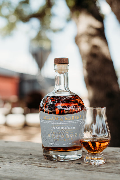 bourbon bottle with a grey label and a glass of poured whiskey in a glencairn in front of the milam and greene distillery blurred in the background